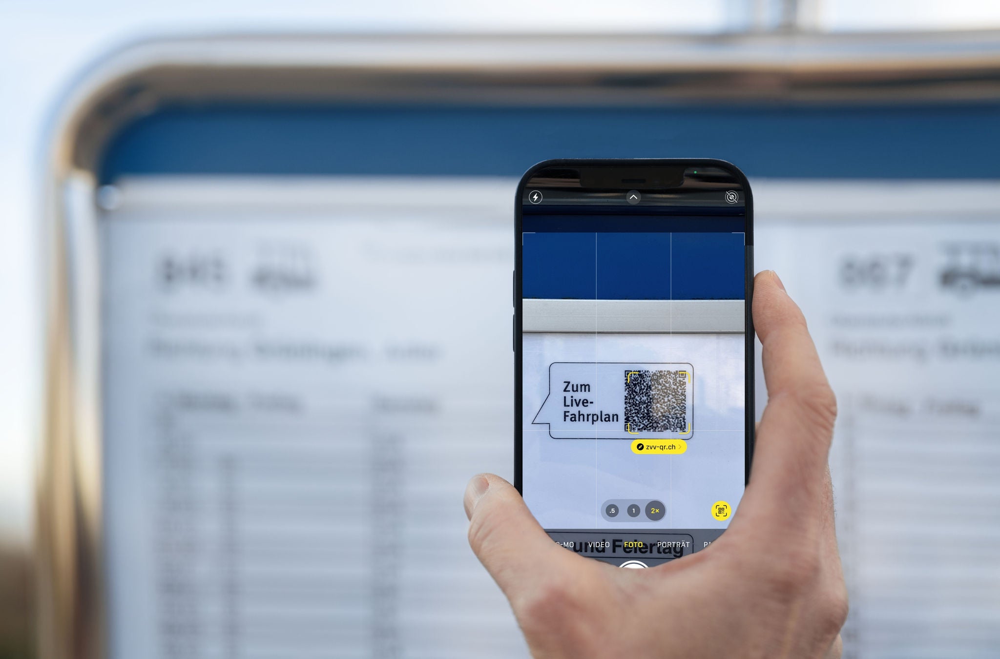 Ein Smartphone wird verwendet, um einen QR-Code zu scannen, der auf einem blauen Fahrplan angebracht ist, um Fahrplaninformationen zu erhalten. Die Szene spielt sich im Freien ab, und der Hintergrund ist unscharf, was den Fokus auf die Handlung des Scannens legt.