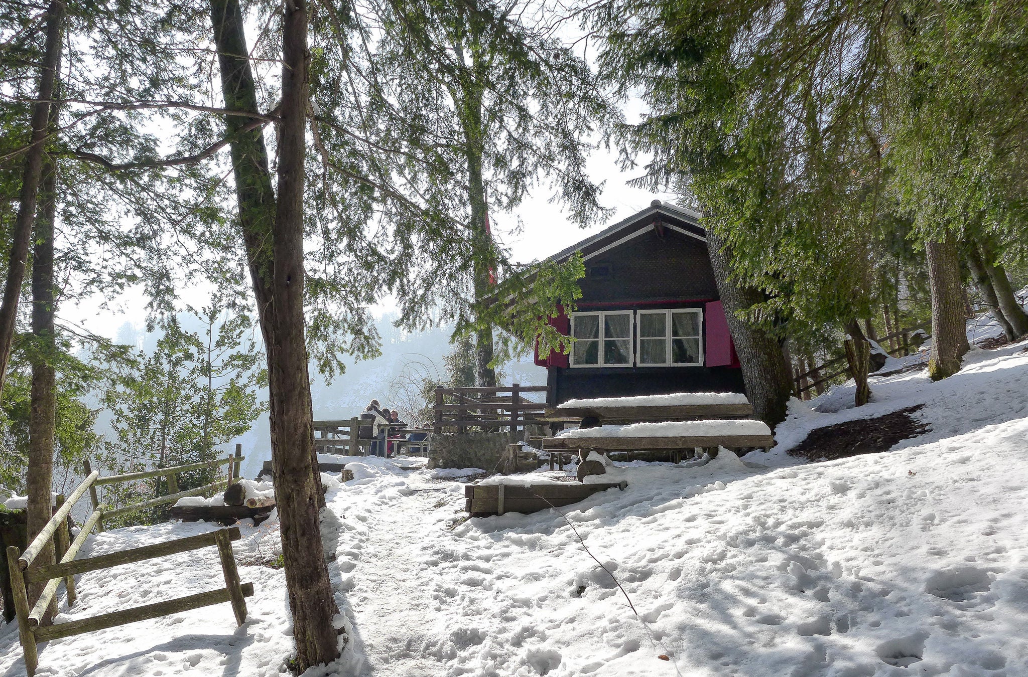 Eine kleine verschneite Hütte mittem im Wald mit Besucher auf der Terrasse